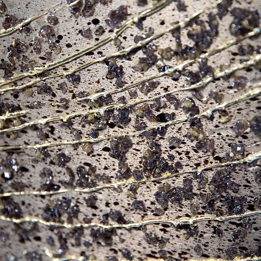 A close-up of a textured metallic surface with horizontal gold-toned lines and scattered dark, faceted crystal-like specks.